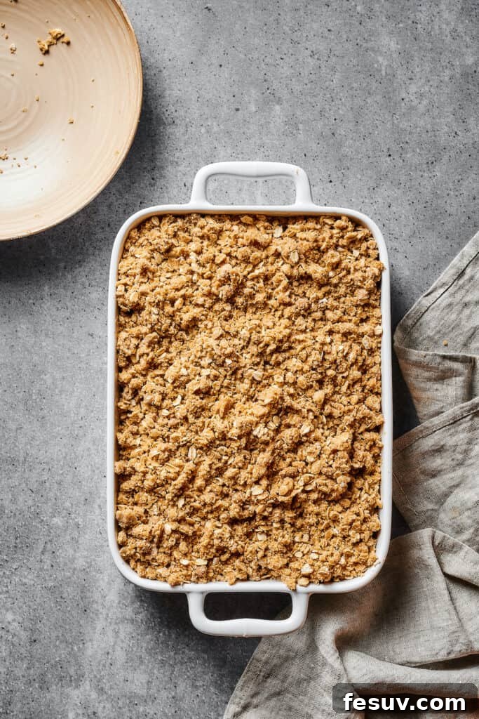 An unbaked apple crisp, with a uniform layer of oat topping over the apple filling, resting in a white casserole dish before going into the oven.