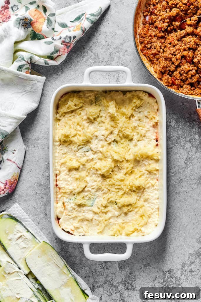 A generous sprinkling of shredded mozzarella cheese over layers of zucchini and cheese filling in a baking dish.