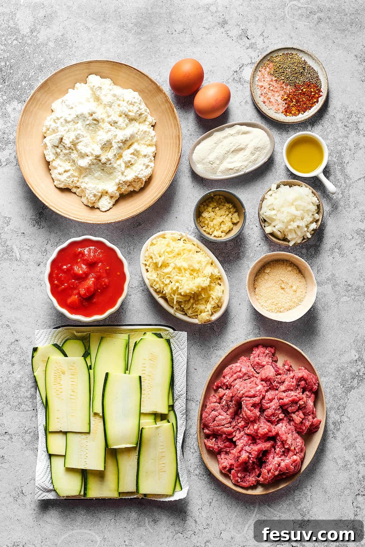 An array of fresh ingredients for zucchini lasagna, including cottage cheese, eggs, seasonings, flour, olive oil, garlic, diced onion, crushed tomatoes, mozzarella, Parmesan, zucchini slices, and ground beef, arranged on a surface.