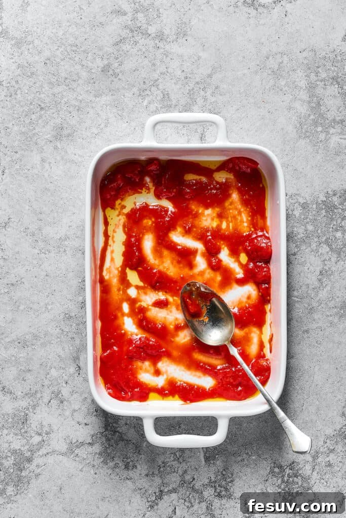 A layer of vibrant crushed tomato sauce evenly spread across the bottom of a white baking dish, ready for lasagna layers.