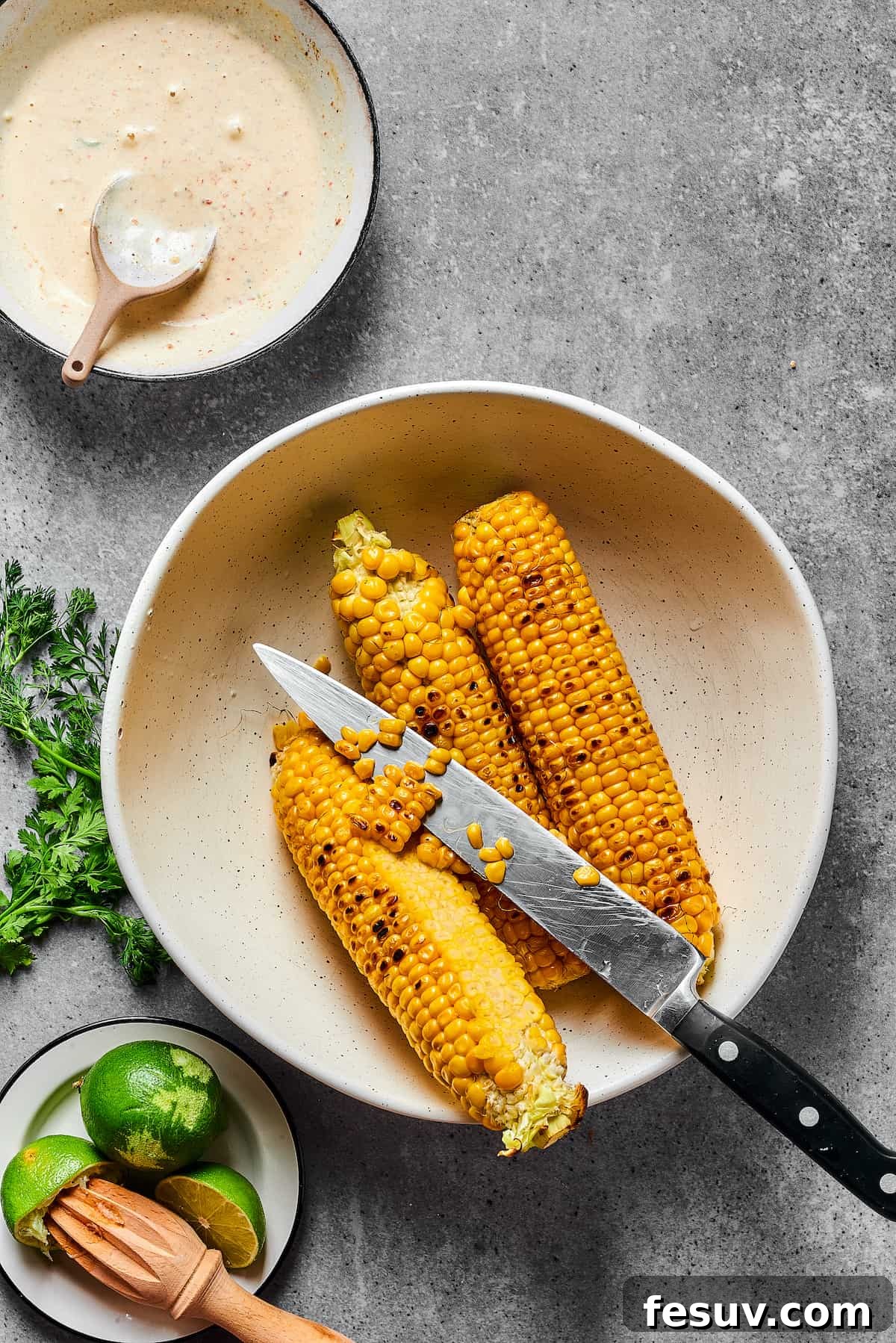 Mexican Street Corn Salad is a mouthwatering dish that’ll have everyone begging for the recipe! Fresh corn kernels in a killer creamy sauce. Grilled corn on the cob with some kernels sliced off into a bowl, next to a knife.