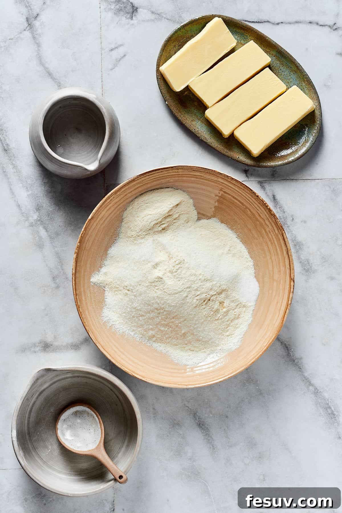 A mixing bowl with flour, sugar, and salt, surrounded by dishes of diced cold butter and ice water.