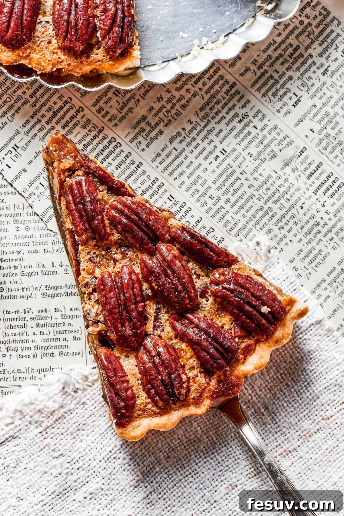 The Best Pecan Pie Recipe is here, easy to make and perfect for your holiday table! You'll love the homemade crust and sweet, nutty filling. overhead shot of a slice of pie held over a newspaper, and a pie plate placed above it.