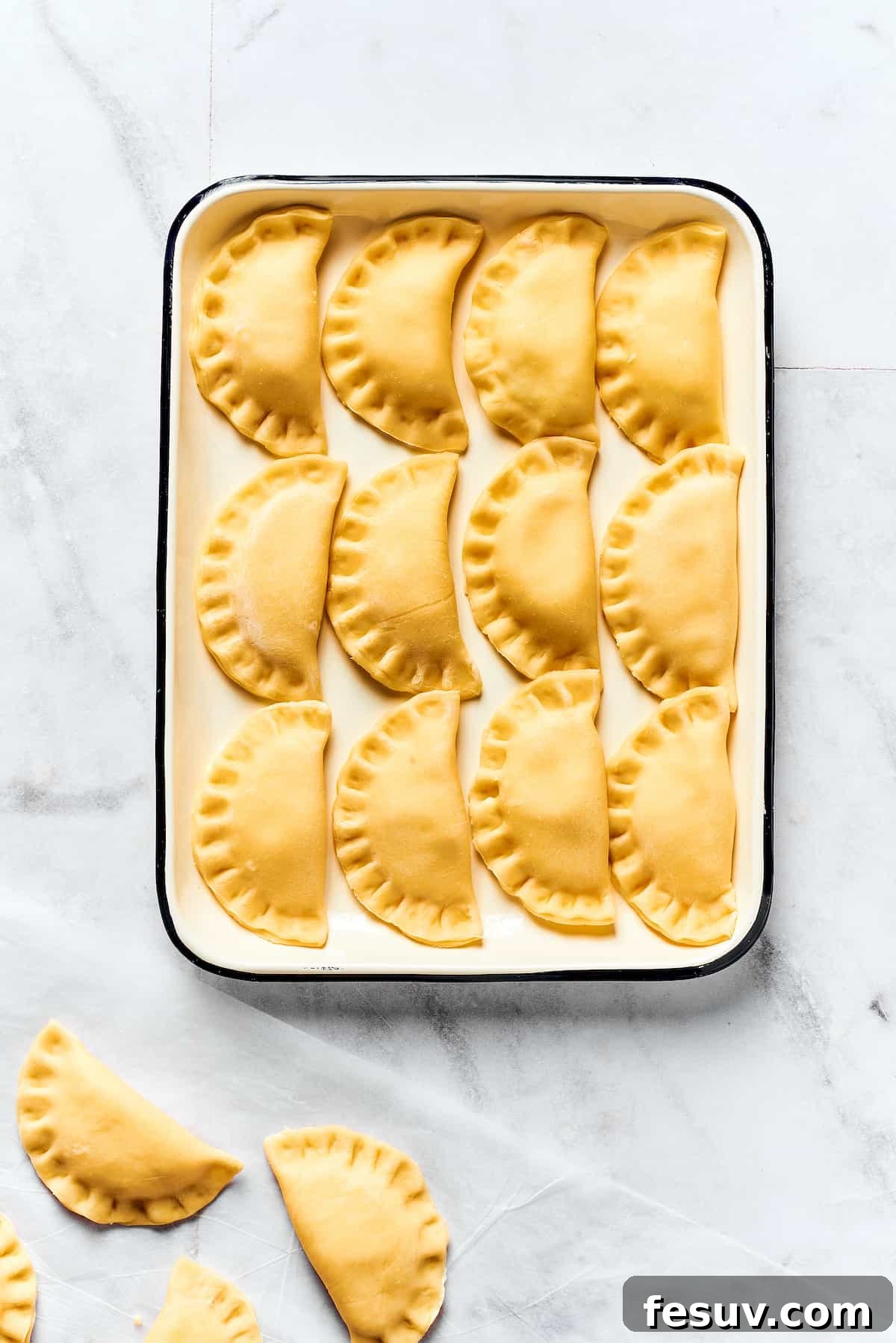 Topped with caramelized onion and crispy bacon, this easy Homemade Pierogi Recipe features a traditional cheese and potato filling. Yum! A tray of uncooked, freshly sealed pierogies lined up on parchment paper, ready for cooking.