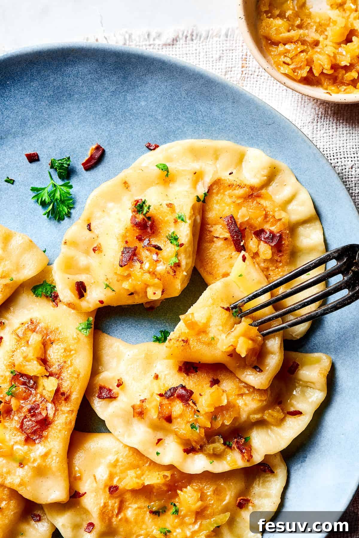 Topped with caramelized onion and crispy bacon, this easy Homemade Pierogi Recipe features a traditional cheese and potato filling. Yum! A fork piercing a bite-sized piece of a golden-brown pierogi, ready to be enjoyed.