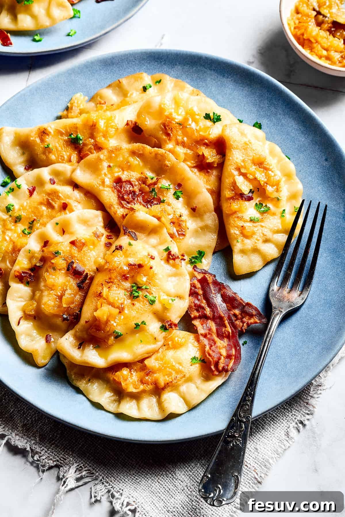 Topped with caramelized onion and crispy bacon, this easy Homemade Pierogi Recipe features a traditional cheese and potato filling. Yum! Homemade potato-filled dumplings served on a blue plate with a fork, ready to be enjoyed.