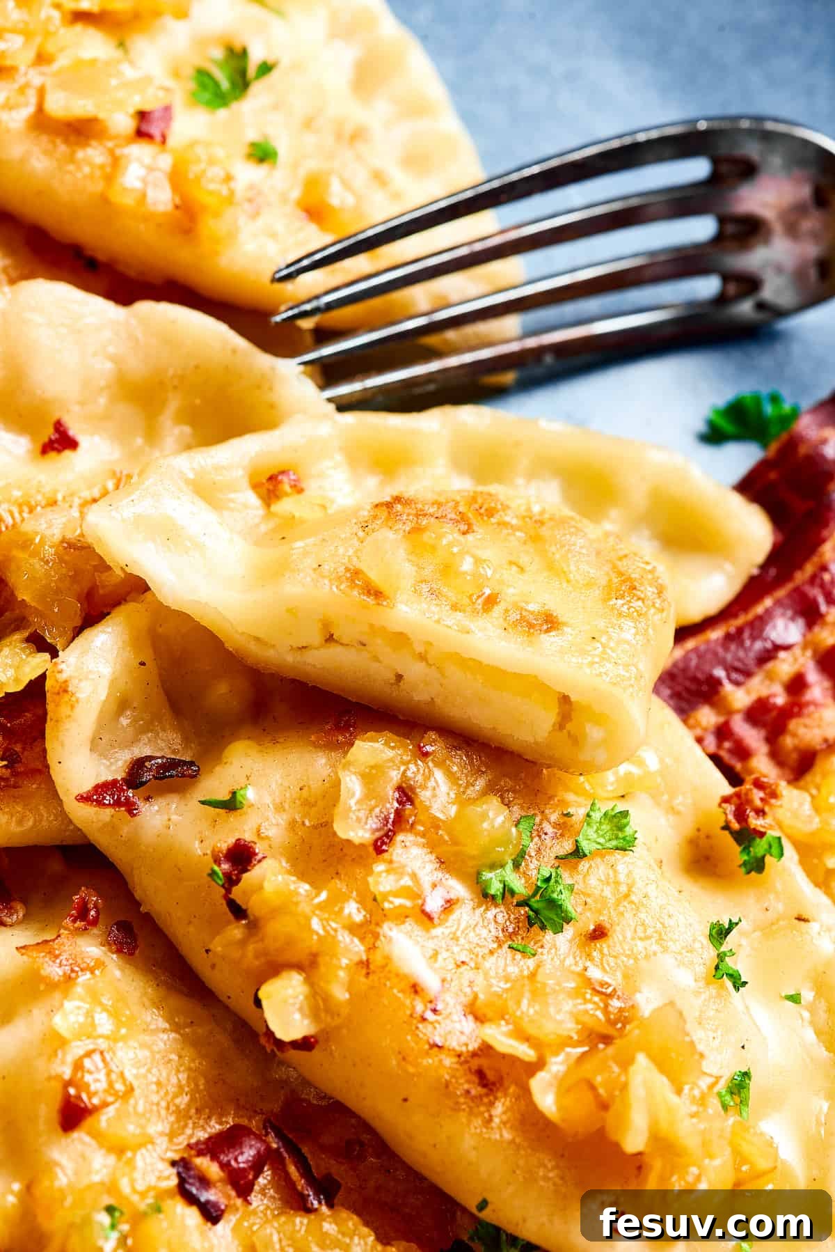 Topped with caramelized onion and crispy bacon, this easy Homemade Pierogi Recipe features a traditional cheese and potato filling. Yum! A plate of traditional homemade pierogies. One has been cut in half with a fork, revealing the creamy potato and cheese filling.