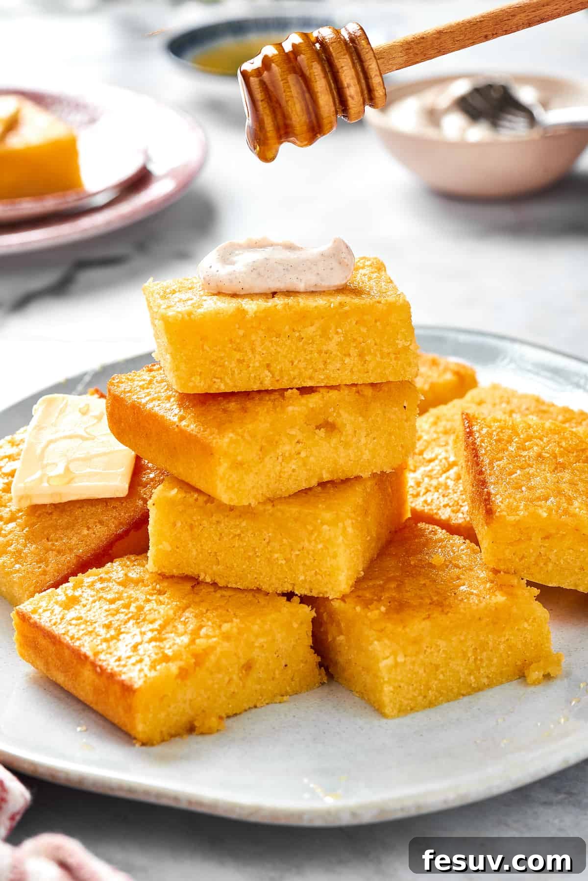A beautiful platter showcases stack after stack of homemade cornbread squares. The top piece is elegantly crowned with a dollop of cinnamon cream, and a honey dipper is poised to drizzle golden honey, highlighting its deliciousness.