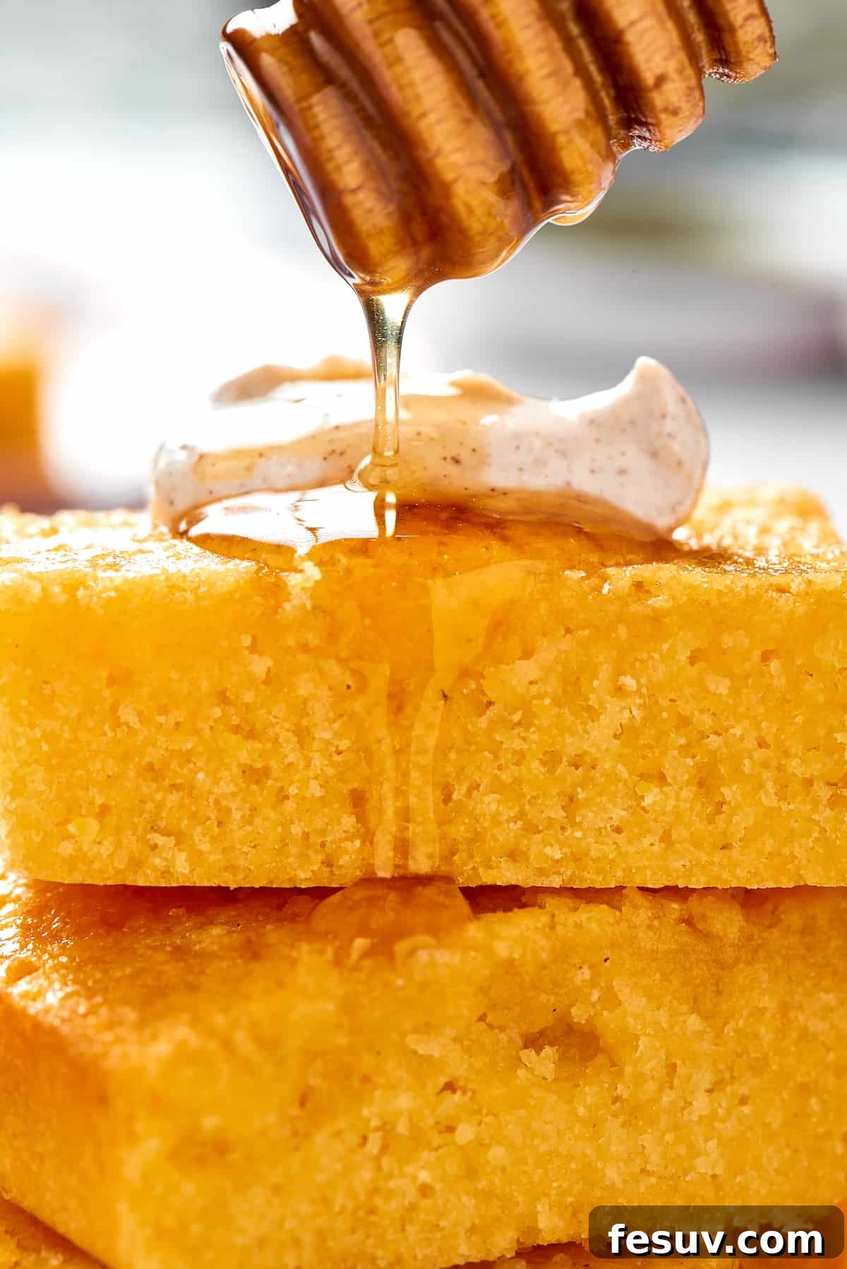 A honey dipper gracefully drizzles glistening honey over freshly cut squares of cornbread, some topped with a dollop of cinnamon cream, highlighting the sweet perfection of this homemade treat.