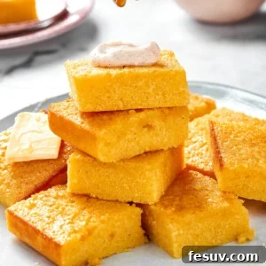 A platter of cornbread with some squares stacked on each other. The top piece of cornbread has a dollop of cinnamon cream on top and is being drizzled with honey.