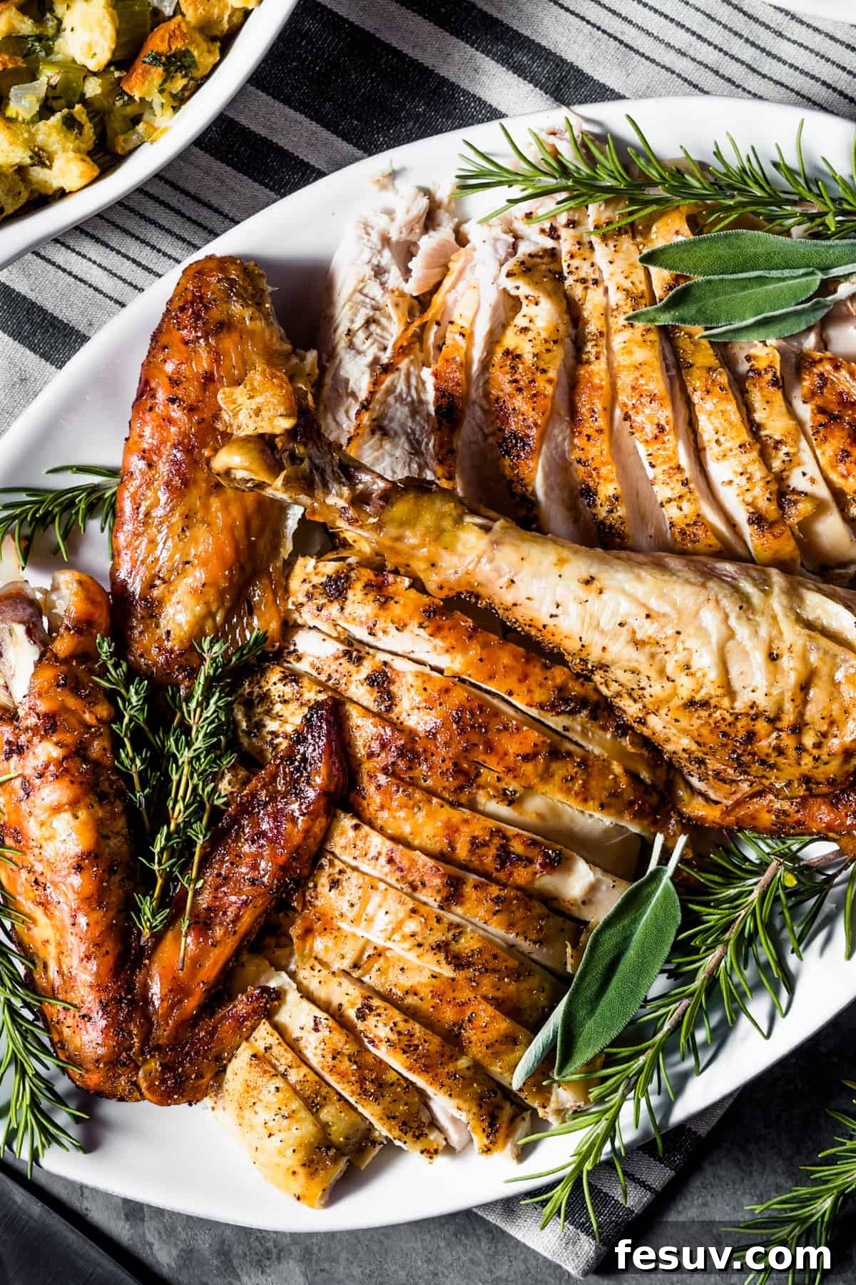 Classic Roast Turkey with Stuffing, fresh herbs, and a pretty pumpkin garnish is easy to make from scratch. The perfect Turkey Day feast! An overhead shot of a perfectly carved roasted turkey, elegantly arranged on a white oval platter and garnished with fresh green herbs, showcasing its golden-brown skin and tender meat.