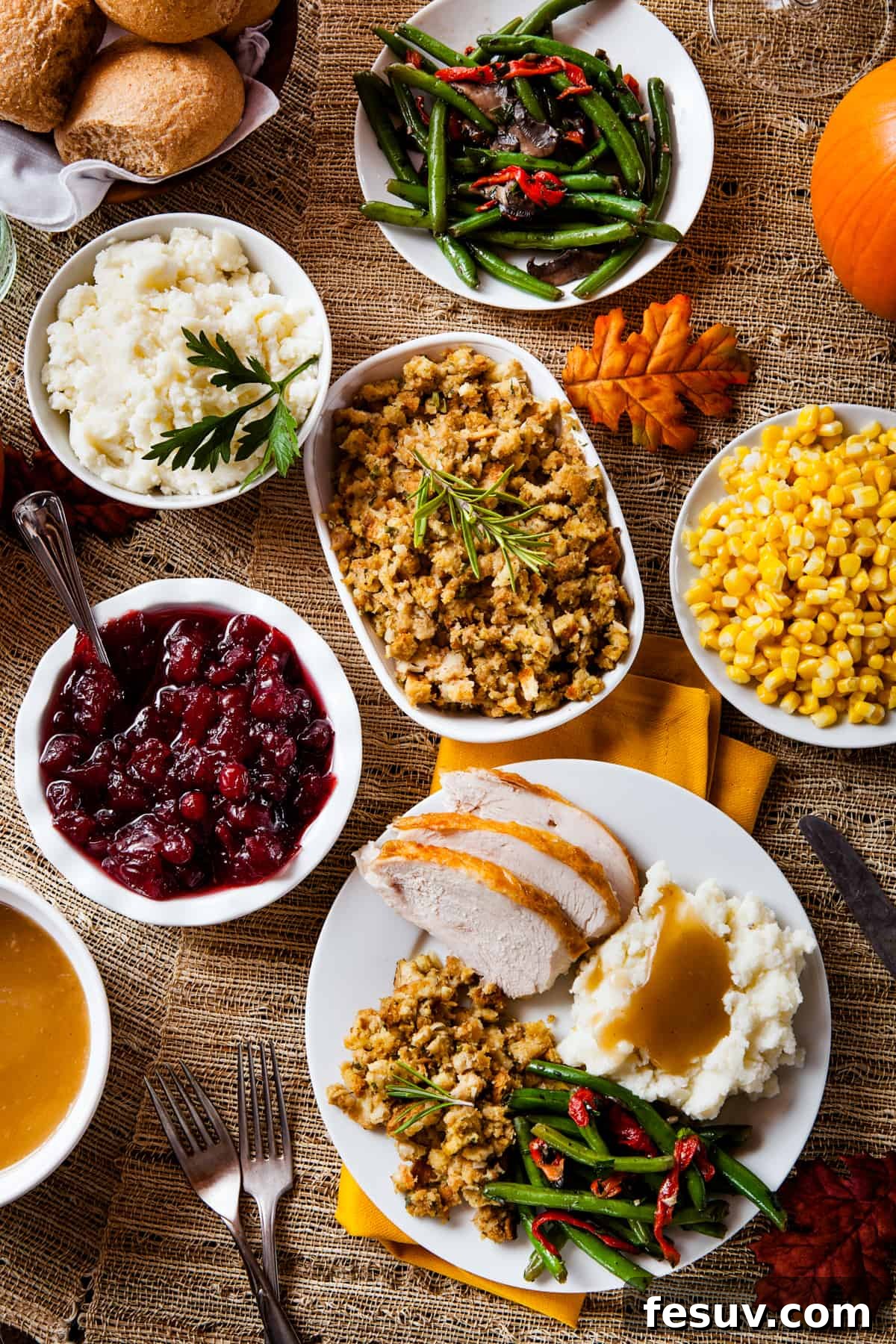 Classic Roast Turkey with Stuffing, fresh herbs, and a pretty pumpkin garnish is easy to make from scratch. The perfect Turkey Day feast! An overhead shot displaying a variety of Thanksgiving side dishes arranged on serving plates. From bottom left: a gravy boat, cranberry sauce in a white bowl, creamy mashed potatoes, savory stuffing, vibrant green beans, sweet corn, and a plate featuring sliced turkey, mashed potatoes, green beans, and corn.