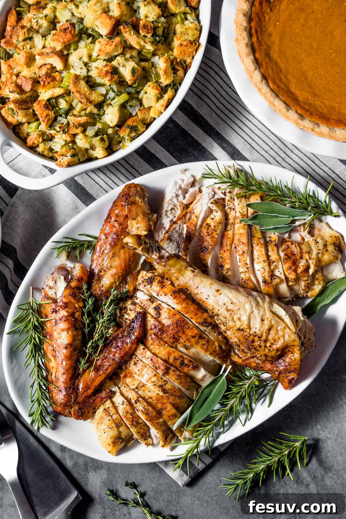 Classic Roast Turkey with Stuffing, fresh herbs, and a pretty pumpkin garnish is easy to make from scratch. The perfect Turkey Day feast! An overhead view of a carved roasted turkey arranged on a white oval platter, garnished with fresh herbs. Adjacent to it are plates of savory stuffing and a slice of pumpkin pie, completing a festive holiday meal.