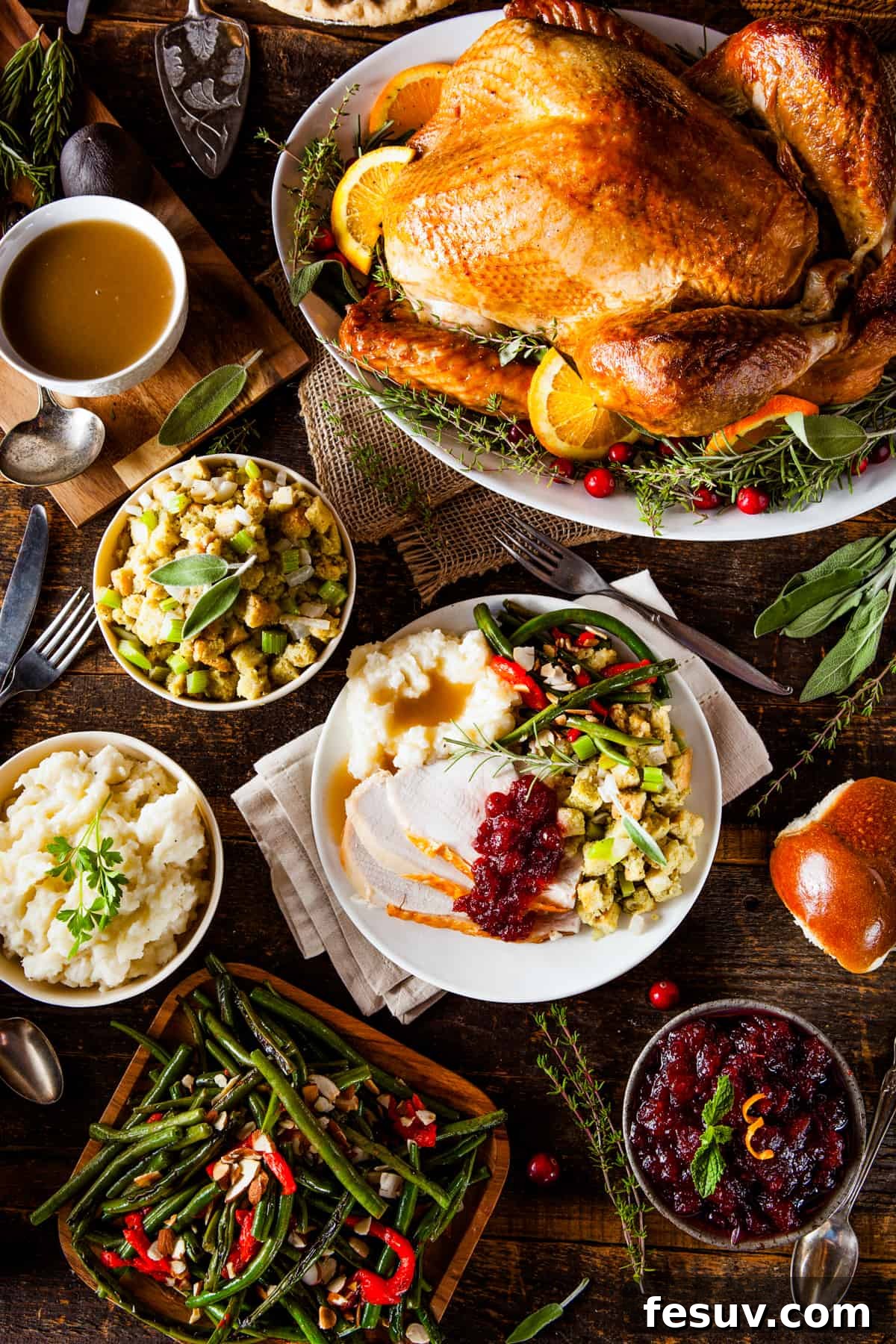 Classic Roast Turkey with Stuffing, fresh herbs, and a pretty pumpkin garnish is easy to make from scratch. The perfect Turkey Day feast! An overhead shot showcasing a whole roasted turkey at the top of the frame, surrounded by an array of delectable side dishes: a bowl of creamy mashed potatoes, a serving of savory stuffing, vibrant green beans, and a bowl of tart cranberry sauce.