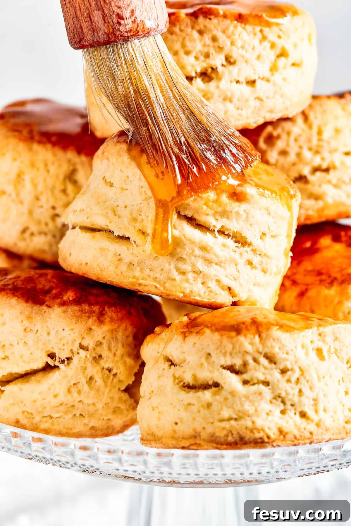 Easy Homemade Biscuits are a buttery, flaky must-have for special meals, and they are so simple to make! Fluffy, melt-in-your-mouth biscuits. A close-up shot of warm, freshly baked biscuits arranged on a baking sheet, with a hand gently brushing maple syrup over their golden tops. This highlights the optional, delicious maple glaze.