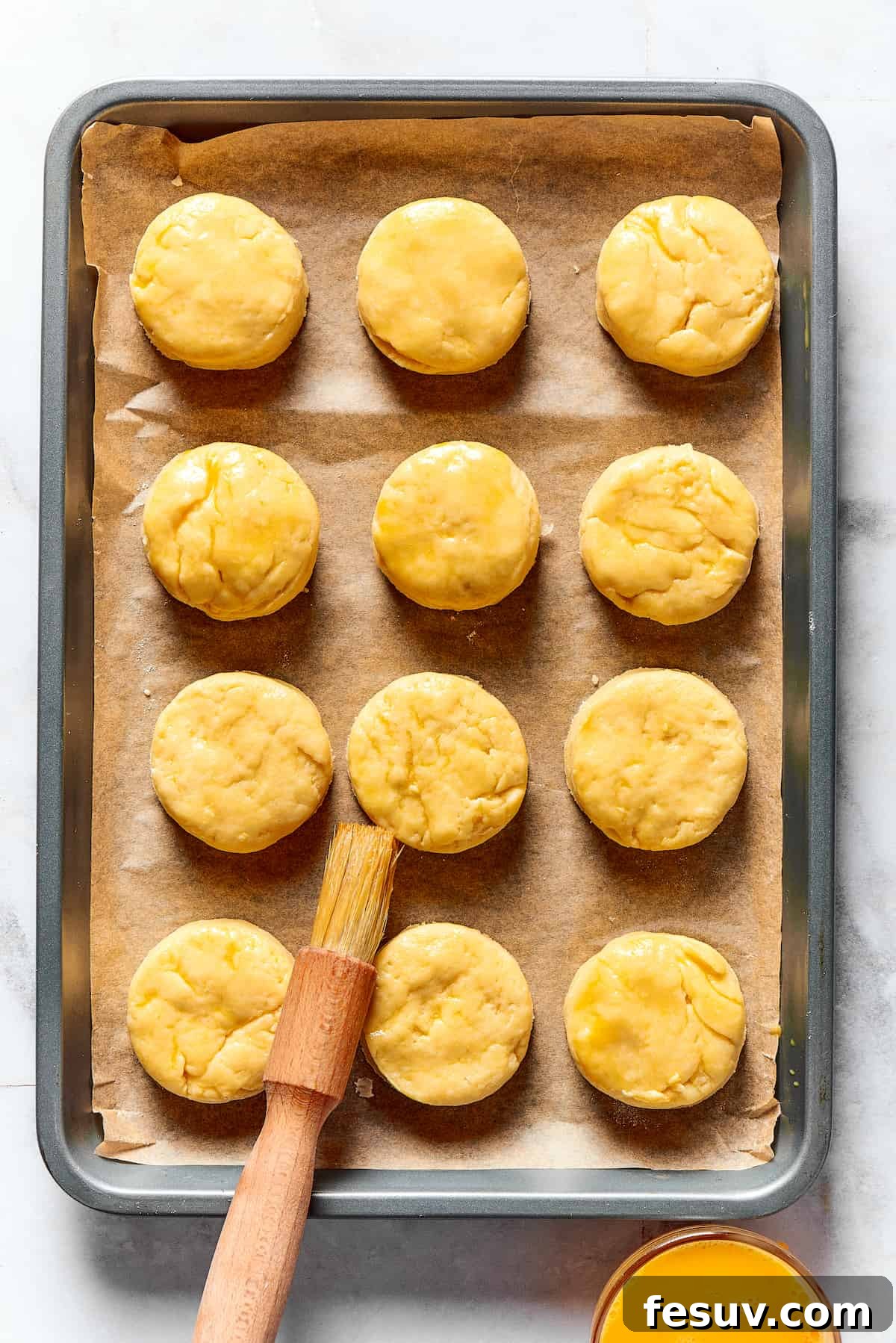 Effortless Fluffy Biscuits 7 A dozen unbaked biscuit rounds are neatly arranged on a parchment-lined baking sheet, being brushed with a beaten egg wash for a glossy finish.