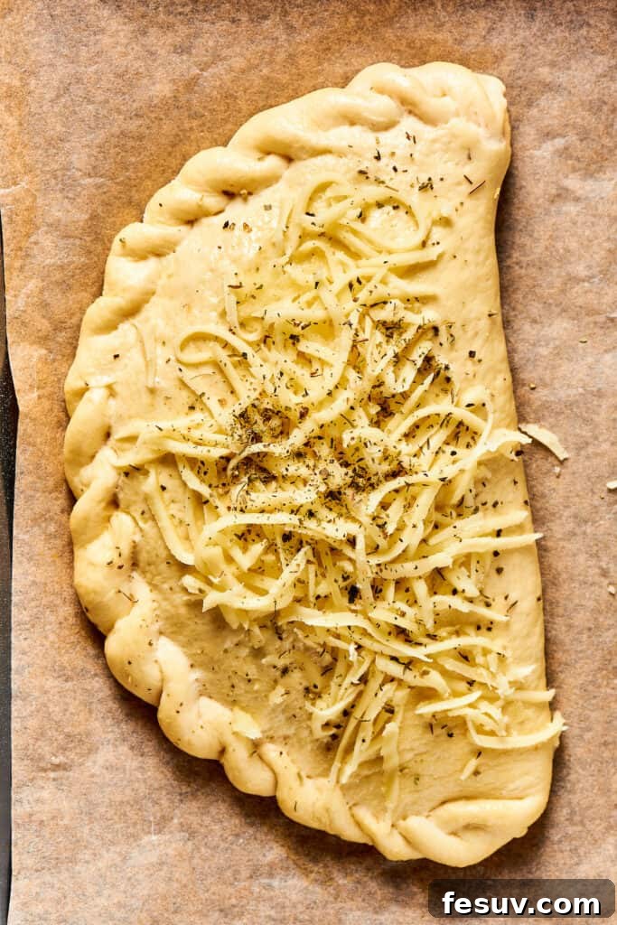 An unbaked calzone, beautifully folded and crimped, topped with a sprinkle of shredded cheese and Italian seasoning.