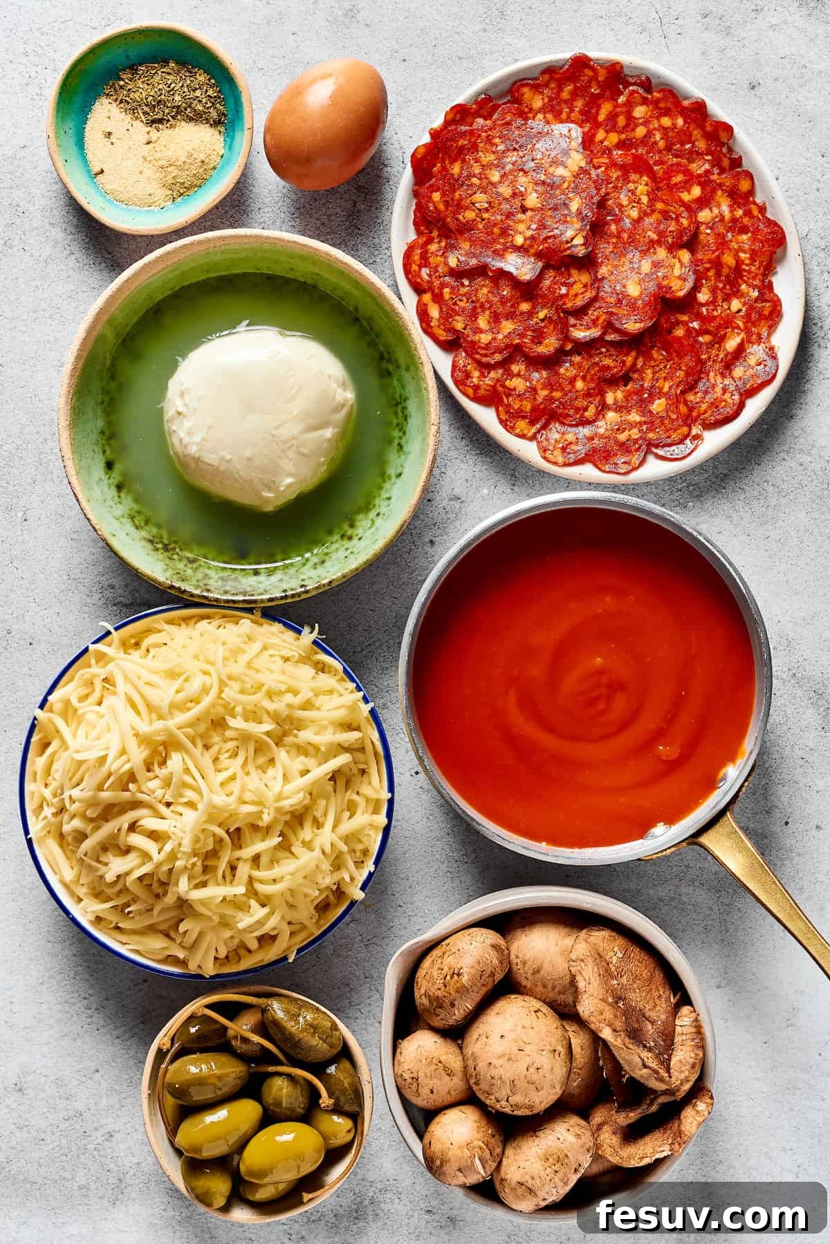 A variety of delicious calzone filling and topping ingredients arranged: Italian seasonings, an egg, pepperoni slices, fresh mozzarella balls, pizza sauce, shredded cheese, olives and capers, and sliced mushrooms.