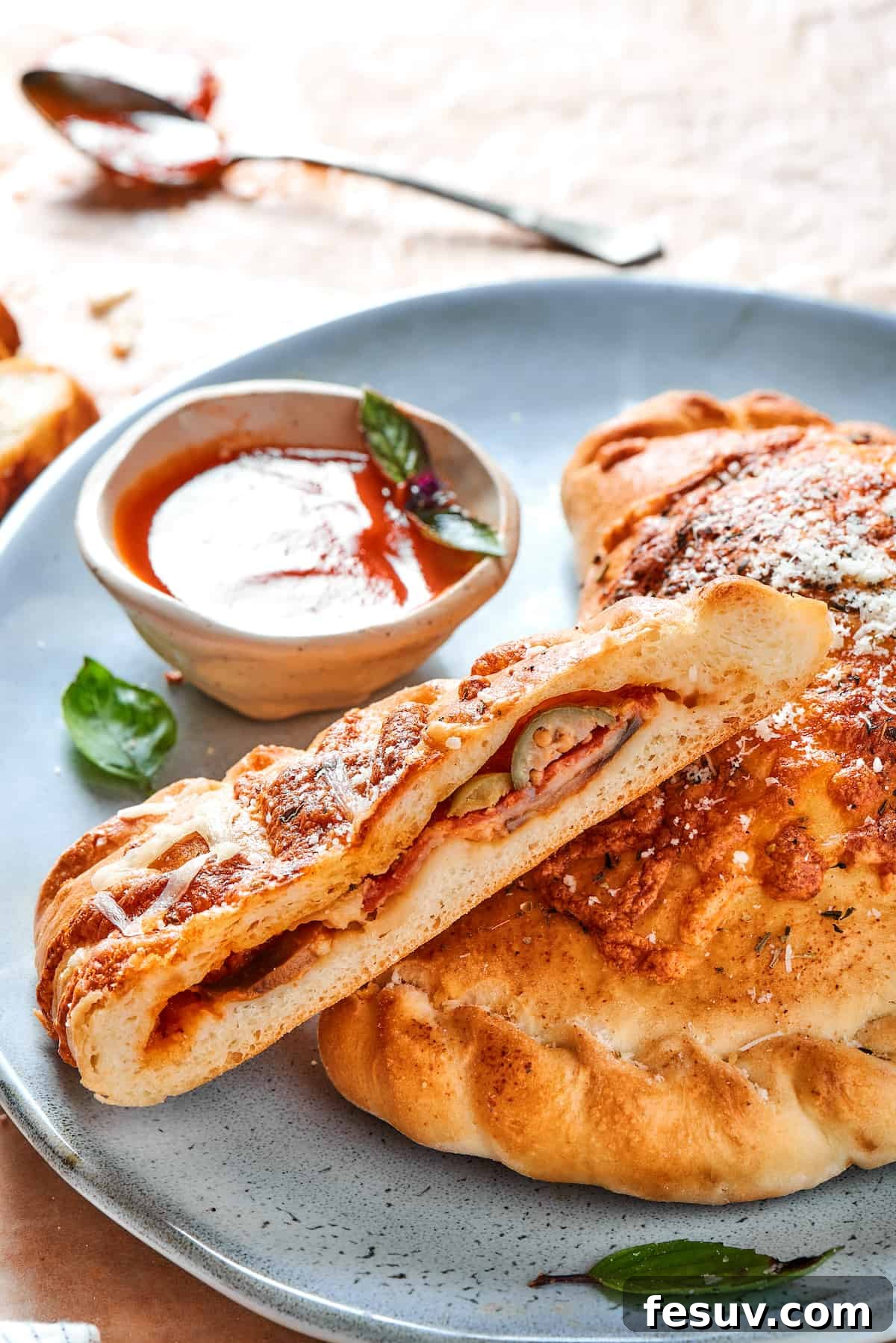 A baked calzone, expertly cut in half, sits on a blue plate alongside a small bowl of rich red dipping sauce, showcasing its delicious, accessible nature.