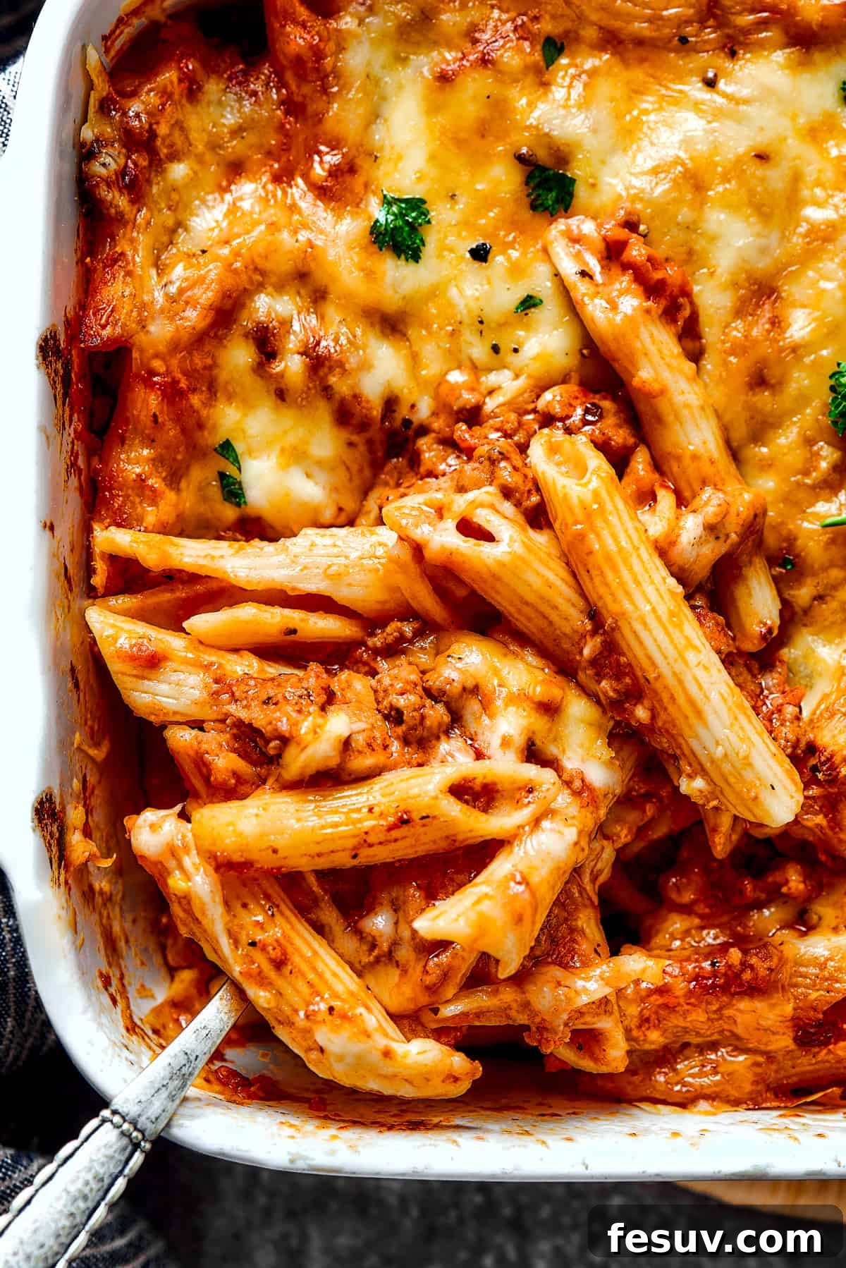 Baked Mostaccioli is a wholesome pasta dinner prepared with a homemade tomato sauce including Italian sausage, ground beef, and lots of cheese! A large baking dish filled with golden-brown, bubbly baked mostaccioli, with a serving spoon scooping out a portion.