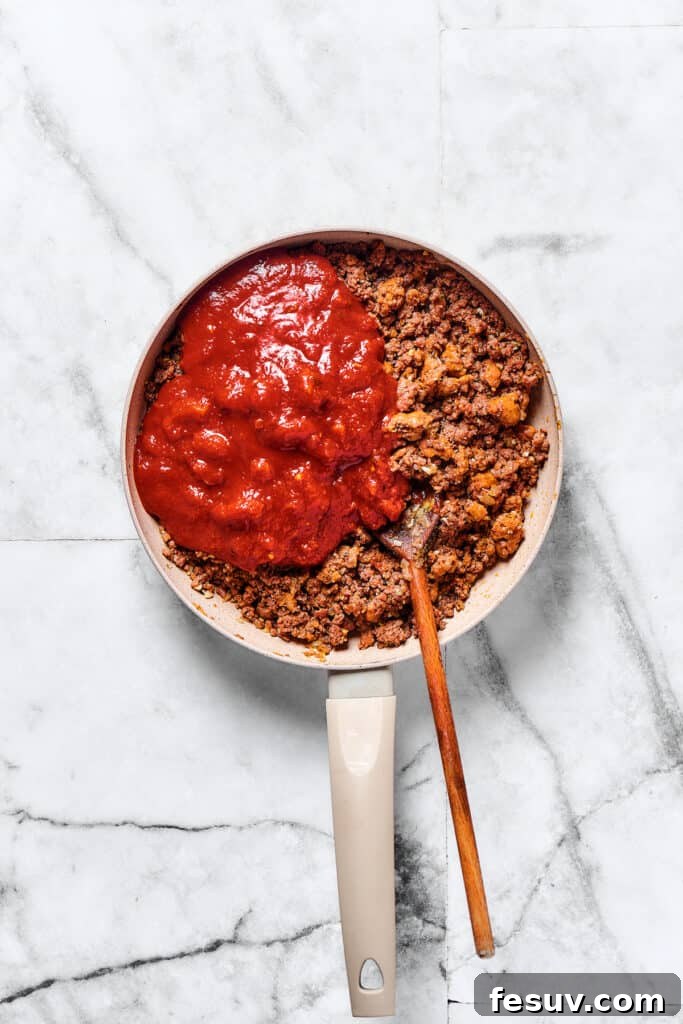 This baked mostaccioli recipe is a hearty pasta dinner prepared with a homemade tomato sauce including Italian sausage, ground beef, and cheese. Crushed tomatoes being stirred into a rich meat sauce in a large skillet, simmering gently.