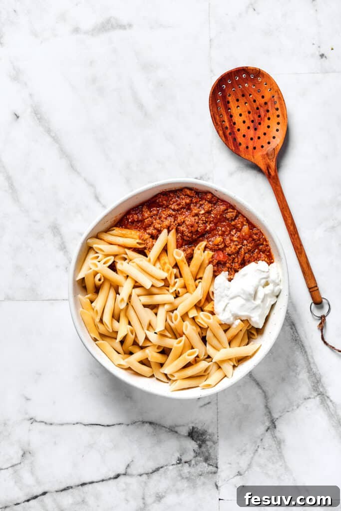 This baked mostaccioli recipe is a hearty pasta dinner prepared with a homemade tomato sauce including Italian sausage, ground beef, and cheese. Mostaccioli pasta, creamy ricotta cheese, and the flavorful meat sauce combined in a large mixing bowl, ready for layering.