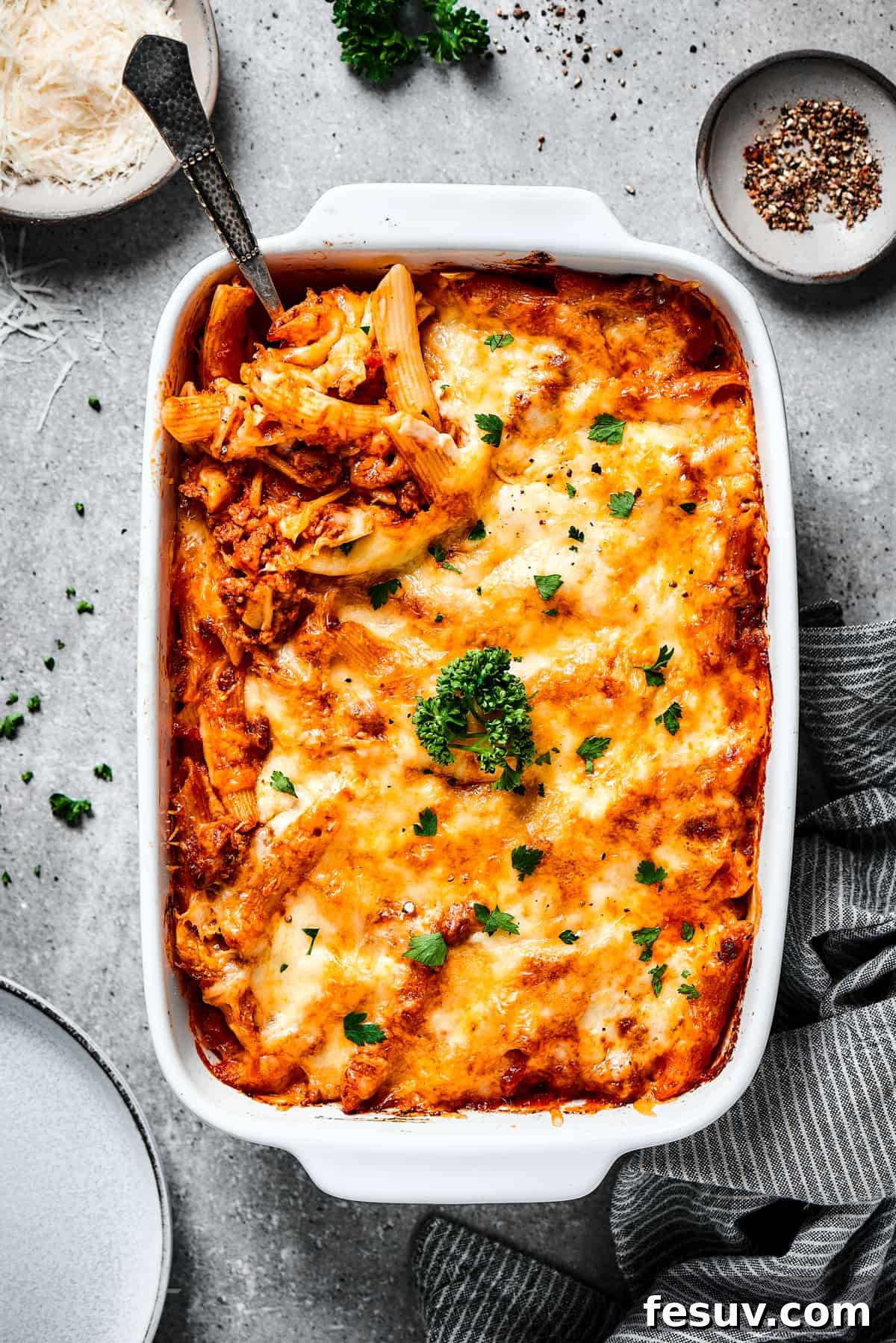 This baked mostaccioli recipe is a hearty pasta dinner prepared with a homemade tomato sauce including Italian sausage, ground beef, and cheese. An overhead shot of a large spoon scooping a generous portion of baked mostaccioli from the baking dish, showing the cheesy, meaty layers.