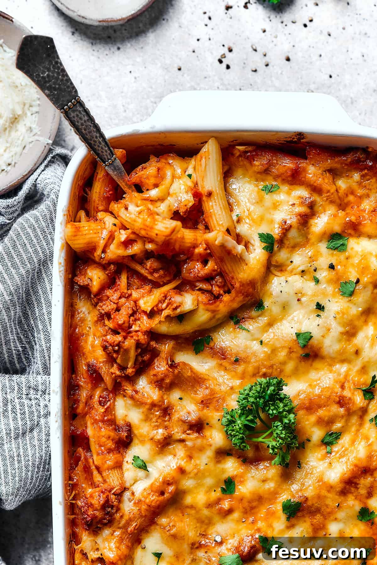 Baked Mostaccioli is a wholesome pasta dinner prepared with a homemade tomato sauce including Italian sausage, ground beef, and lots of cheese! A serving spoon lifting a portion of hot, cheesy baked mostaccioli from a large white baking dish, ready to be enjoyed.