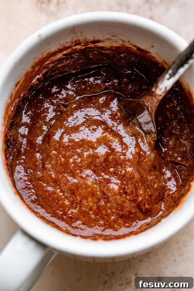 This moist and indulgent Chocolate Mug Cake cooks in one minute inside the microwave. It is the most perfect, individual-sized dessert! A fork stirring through a rich chocolate batter inside a mug, ensuring all ingredients are thoroughly combined for the mug cake.