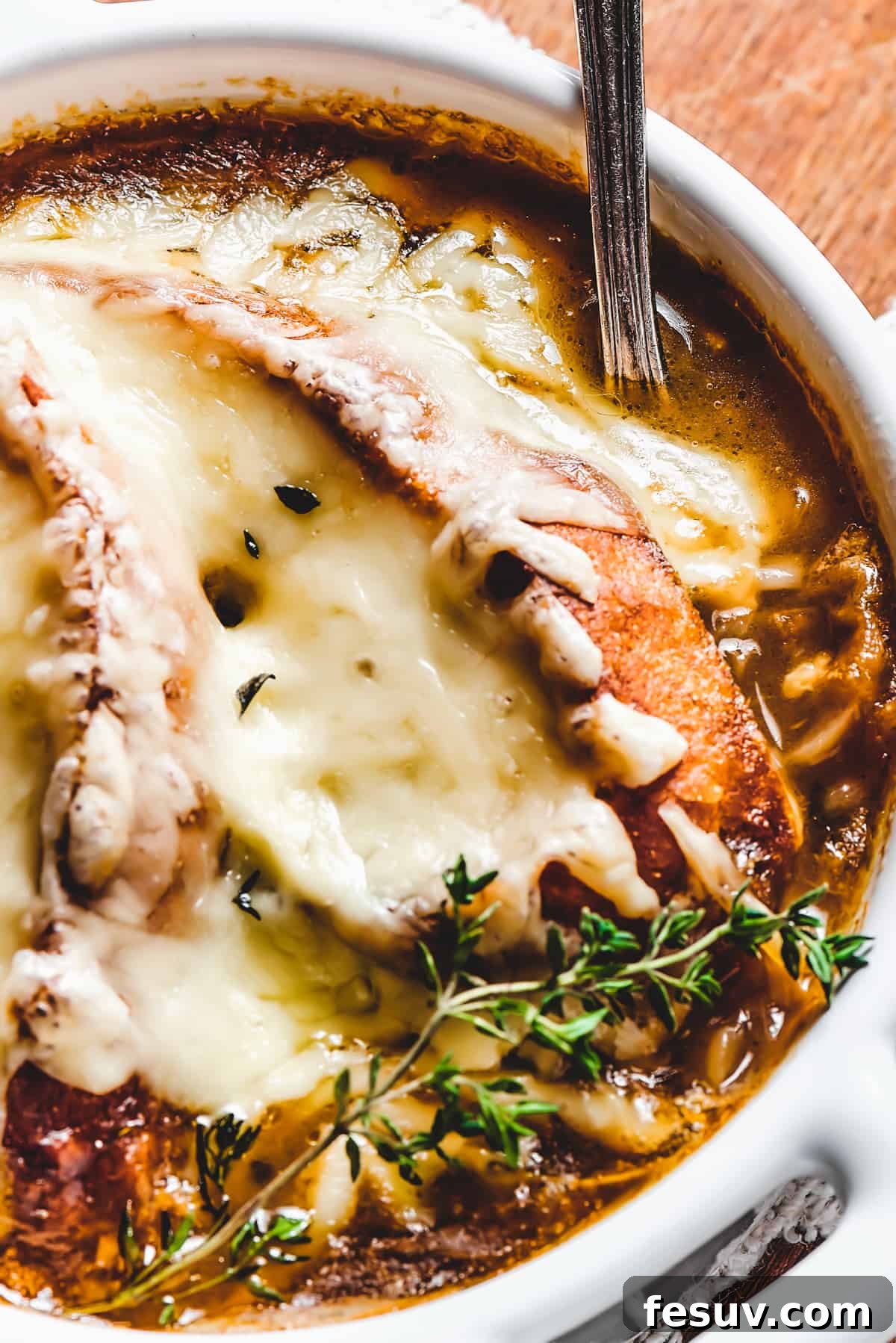 A close-up of French Onion Soup, showcasing the irresistible melted cheese and golden-brown crouton. Close-up shot of a single serving of French Onion Soup, with richly melted cheese draped over a baguette crouton and a sprig of fresh thyme.