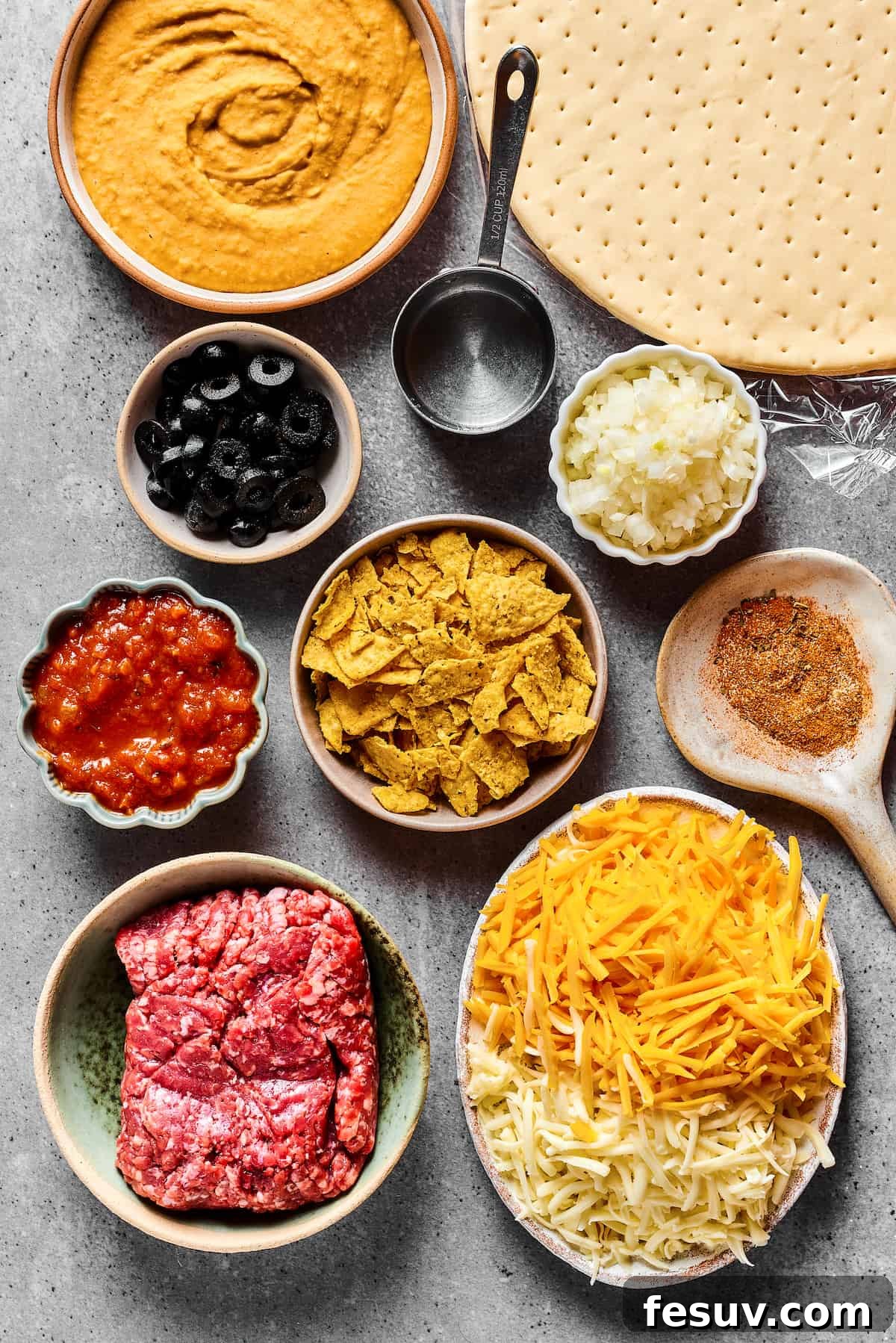 A visual lineup of Taco Pizza ingredients: from top, refried beans, pizza crust, black olives, water, onion, salsa, crushed tortilla chips, taco seasoning, ground beef, and cheese, all ready for assembly.