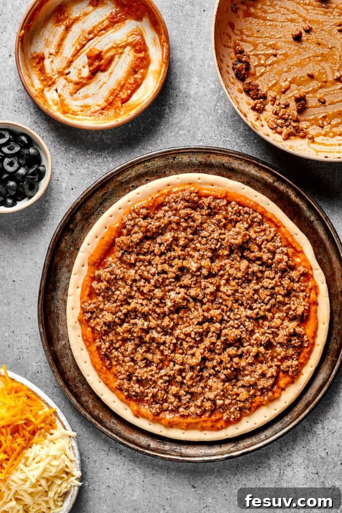 A pizza crust artfully layered with refried beans, salsa, and seasoned taco meat, ready for cheese.