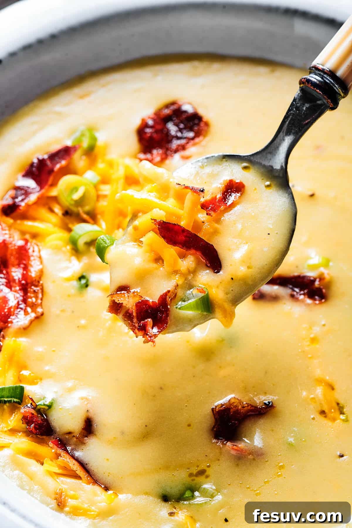Ultimate Cheesy Bacon Potato Soup 12 A pristine white bowl filled with creamy, rich loaded baked potato soup, with a spoon gently lifted, showcasing the perfect texture and tempting toppings.
