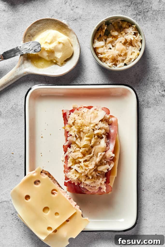 One slice of rye bread, spread with Russian dressing, is generously topped with two slices of Swiss cheese, a pile of savory corned beef, and a layer of tangy sauerkraut. A second slice of bread with dressing and cheese awaits nearby for assembly.