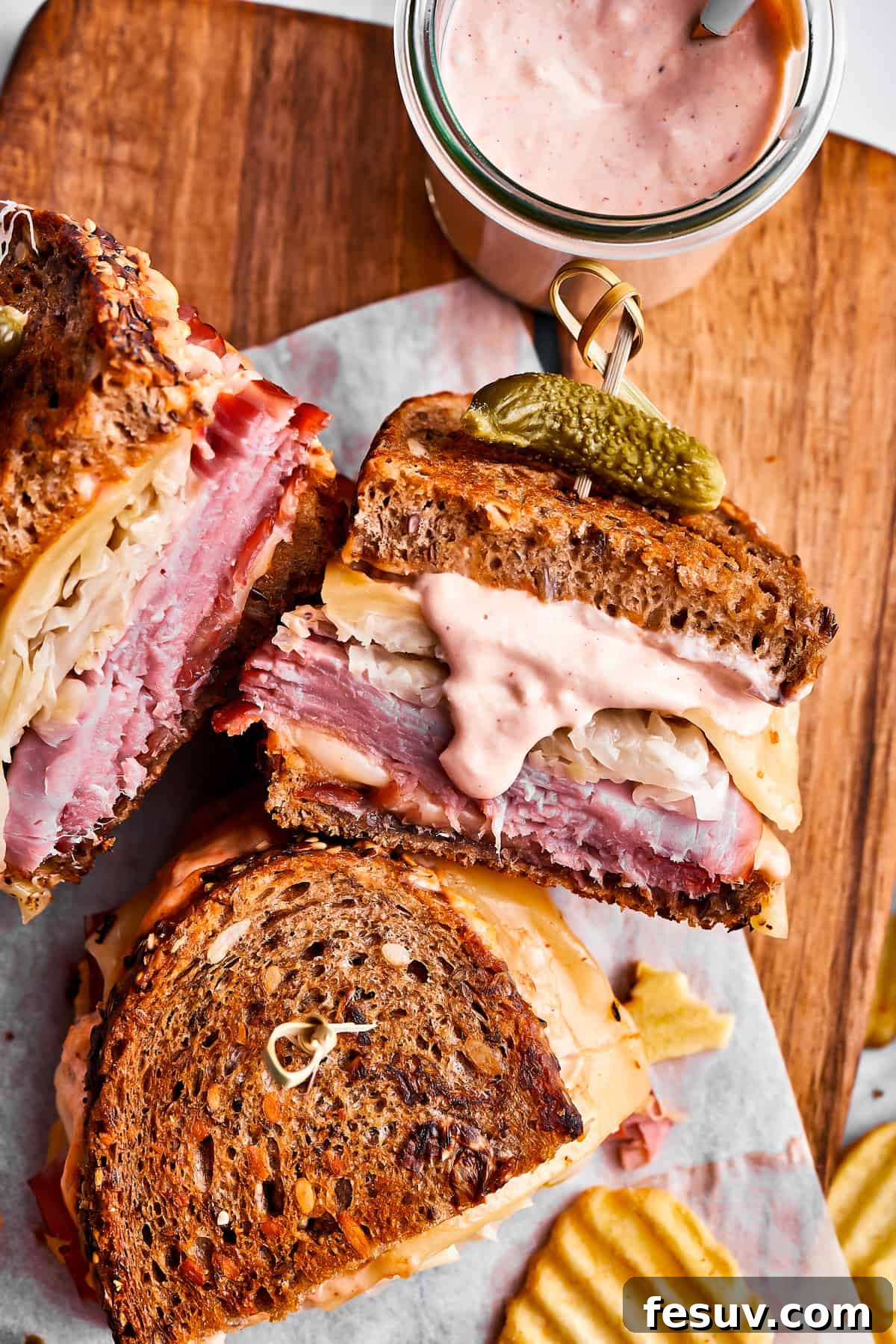 Three expertly prepared Reuben sandwich halves, featuring golden-brown grilled bread and layers of melted cheese, corned beef, and sauerkraut, are arranged on a wooden cutting board alongside a bowl of Russian dressing and a generous serving of crispy potato chips.