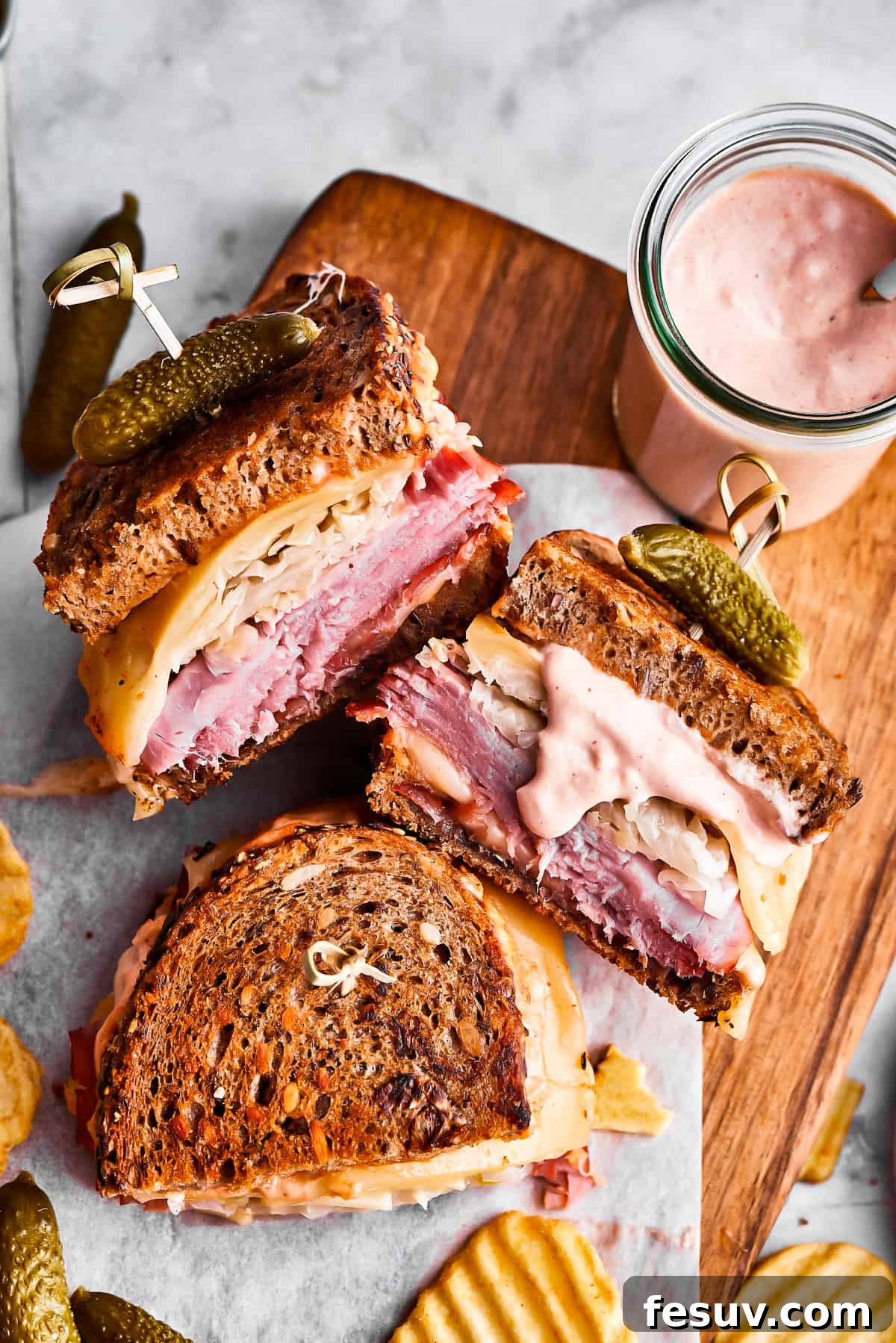 Three perfectly grilled Reuben sandwich halves, featuring melted Swiss cheese and vibrant fillings, are artfully arranged on a cutting board alongside a generous portion of crispy potato chips and a jar of creamy Russian dressing.