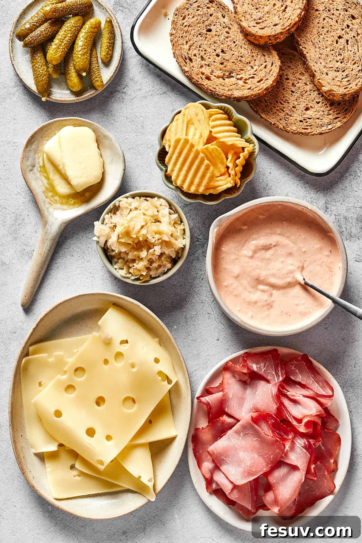 All the vibrant, fresh ingredients needed for a perfect Reuben sandwich are meticulously arranged on a rustic wooden surface: crisp pickles, slices of hearty rye bread, crunchy potato chips, a dollop of creamy Russian dressing, thinly sliced corned beef, generous slices of Swiss cheese, well-drained sauerkraut, and softened butter.