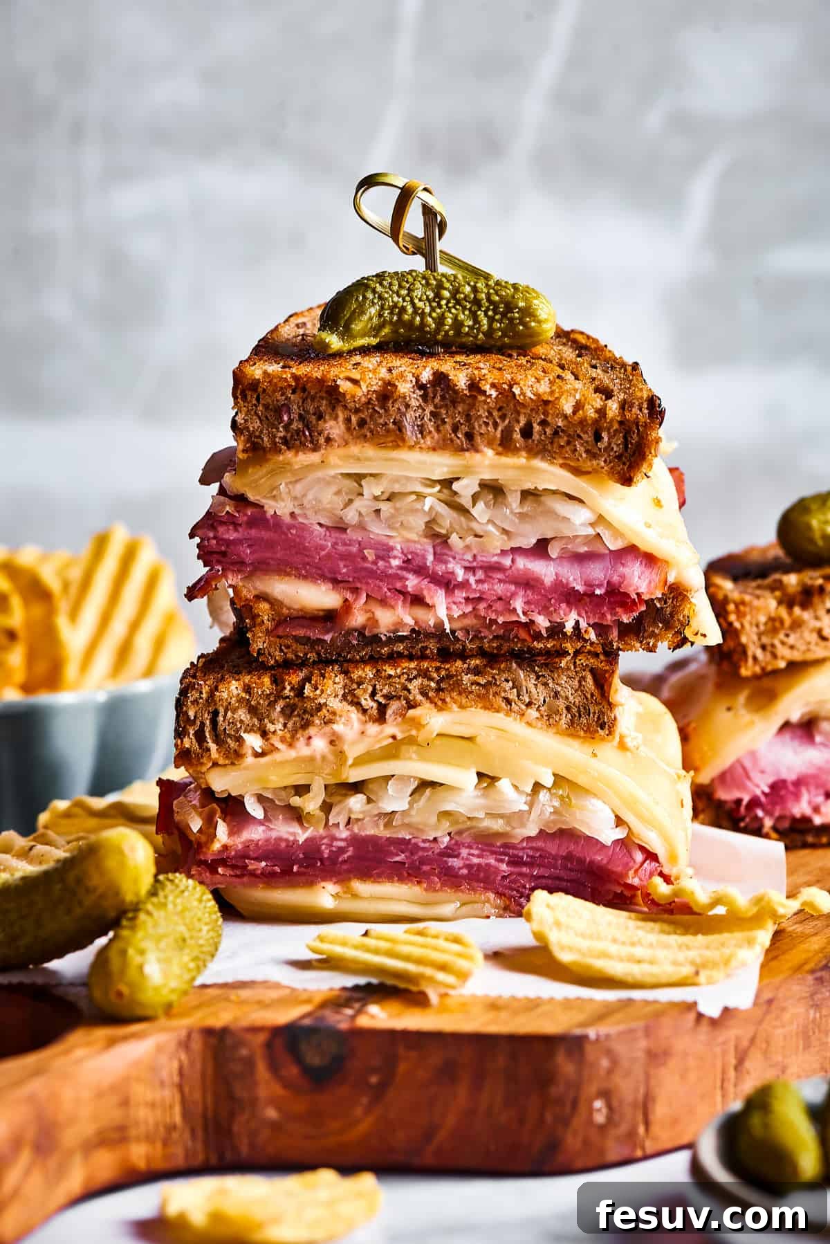 A stunning spread of freshly prepared Reuben sandwiches, expertly cut in half to reveal their savory layers, accompanied by a bowl of creamy Russian dressing, a pile of golden potato chips, and crisp dill pickles, all arranged on a rustic wooden cutting board.