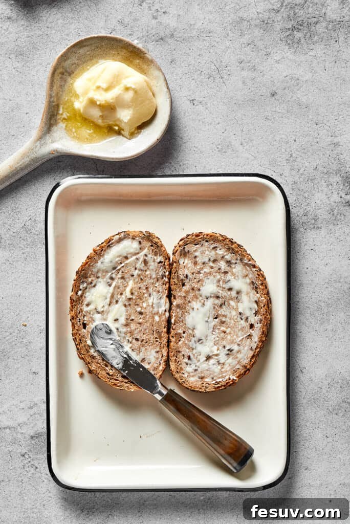 A hand expertly spreading a thin, even layer of softened butter across one side of a slice of rye bread, preparing it for the perfect golden-brown grill.