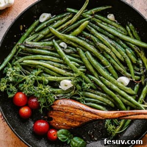 green beans, tomatoes, and garlic cloves in a black skillet with a wooden spoon inside