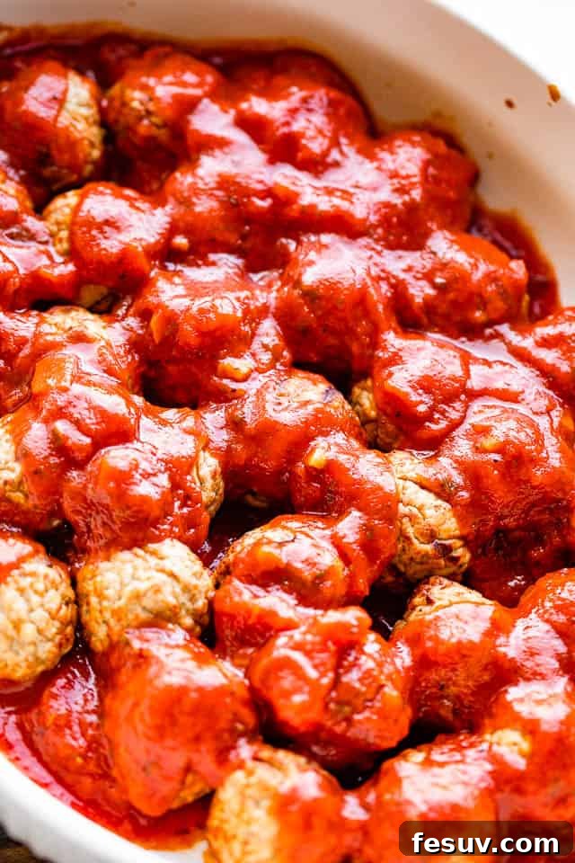Pouring rich marinara sauce generously over a bed of perfectly baked mini meatballs arranged in a white casserole dish, preparing for the final cheesy bake.