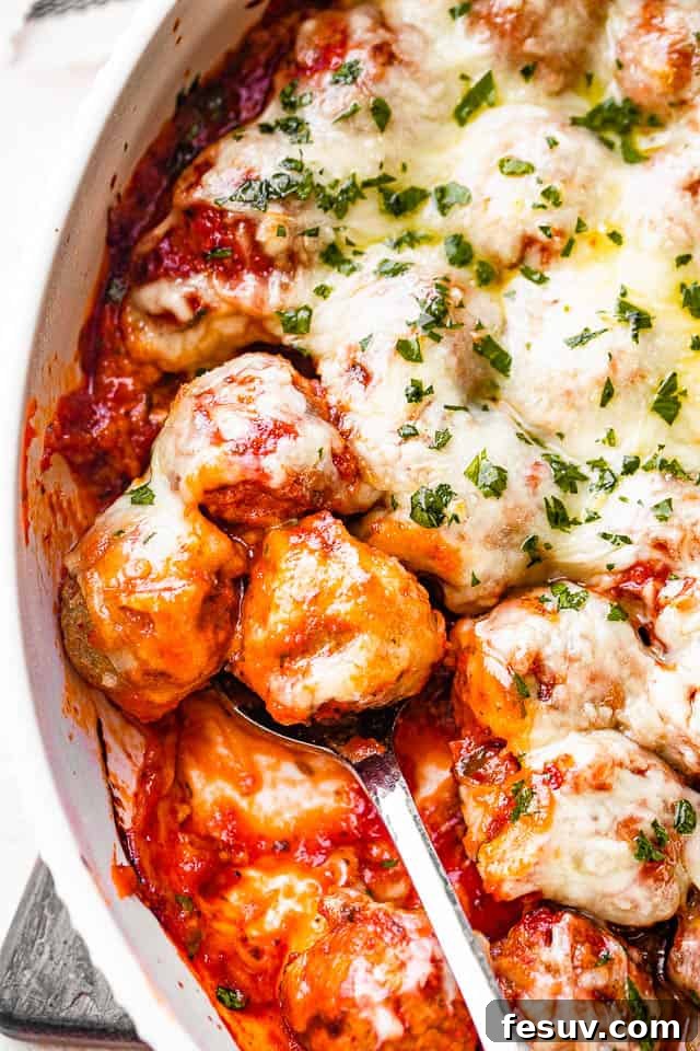 A serving spoon lifting a portion of cheesy baked meatballs, rich with marinara and melted mozzarella, from a white baking dish, ready to be plated.