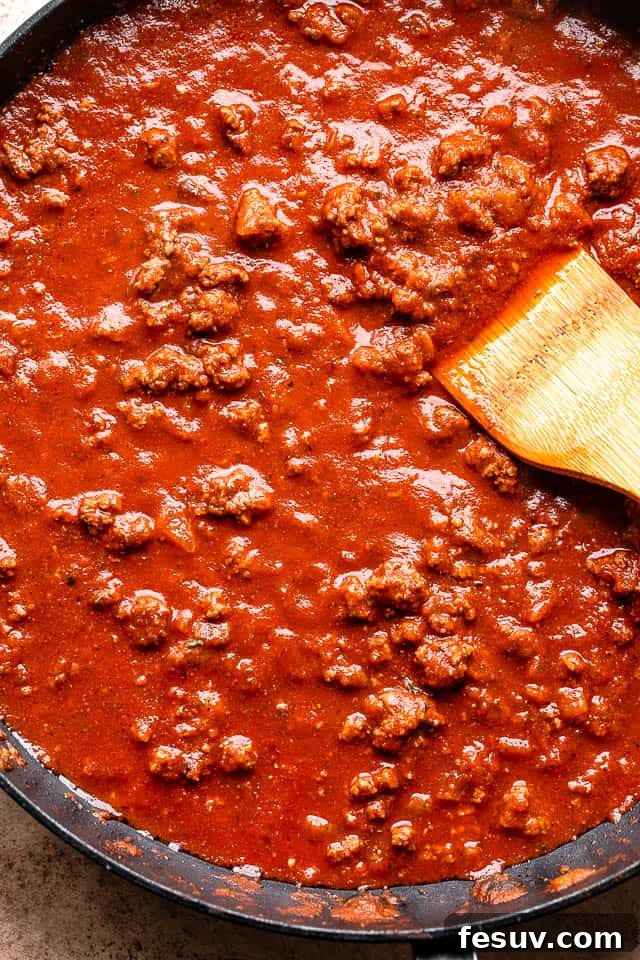 The process of making a rich tomato sauce with ground beef, simmering gently in a black skillet on the stovetop, filling the kitchen with aromatic spices.