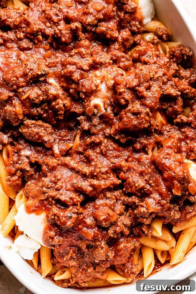 Rich ground beef tomato sauce being generously poured over a bed of perfectly cooked penne pasta, preparing for the cheesy baked ziti layering.