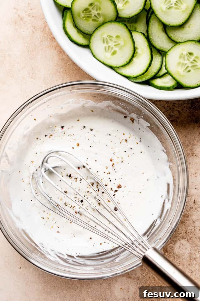 A pristine glass bowl holding the creamy sour cream dressing, with a whisk resting inside, ready to be poured over the cucumbers.