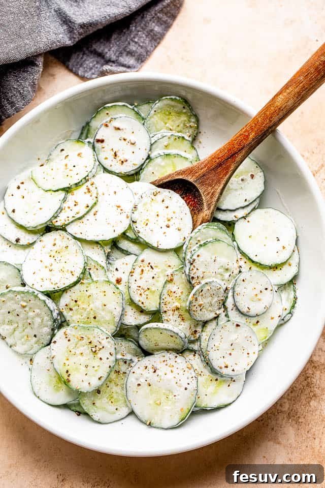 A wooden spoon gently mixing fresh cucumber slices with creamy sour cream dressing in a white ceramic salad bowl.