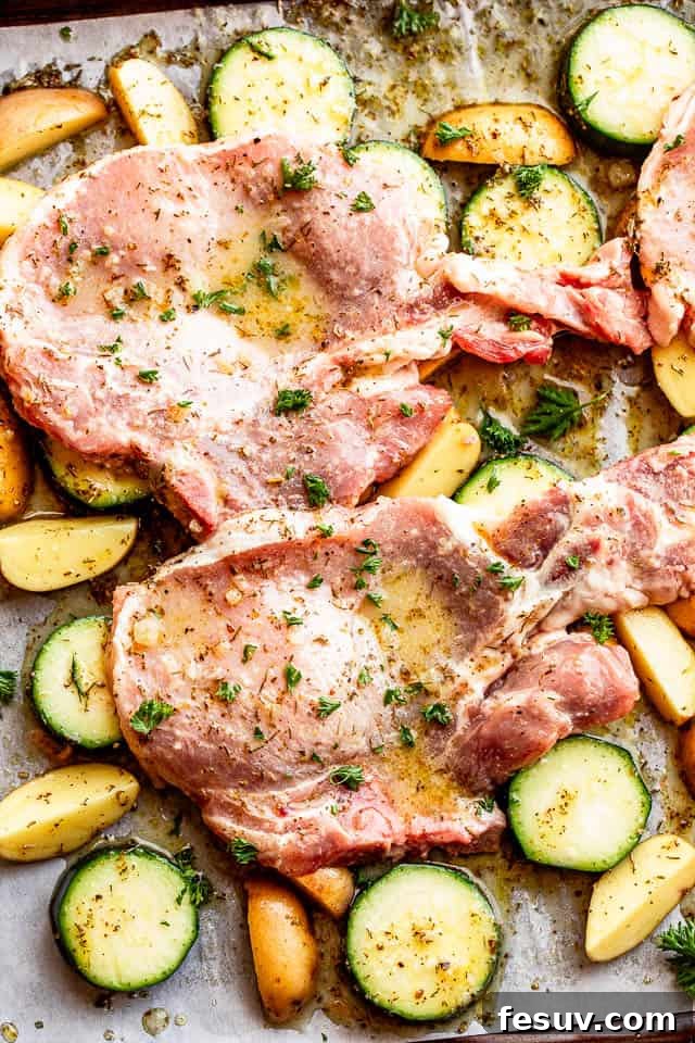 A baking sheet featuring raw, marinated pork chops nestled among slices of zucchini and quartered baby potatoes, ready for the oven.