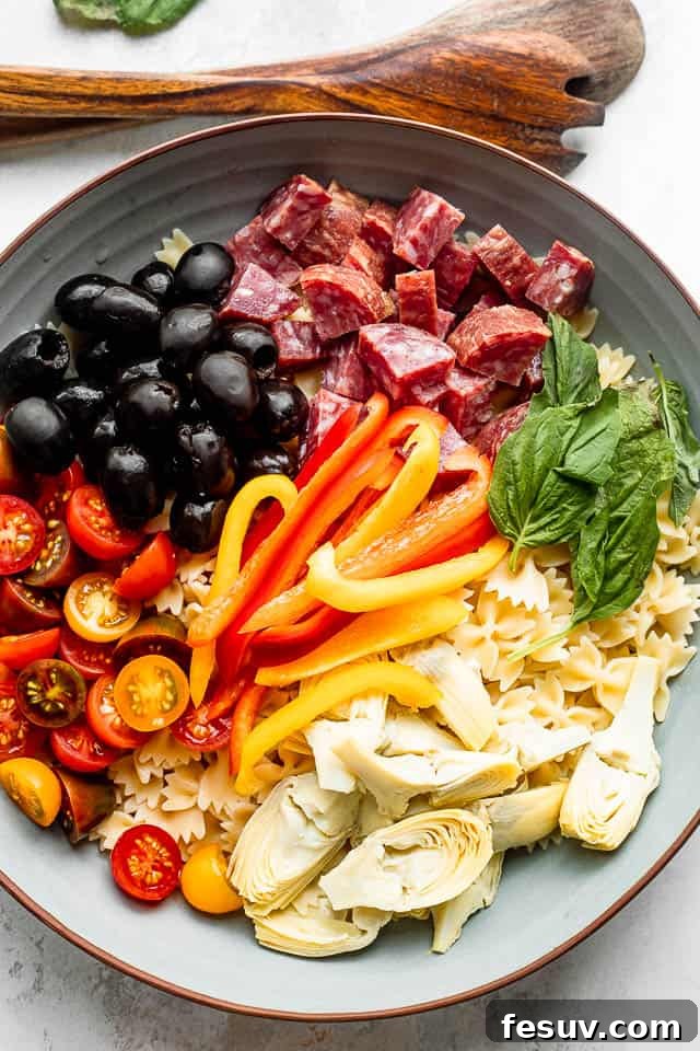 Colorful Antipasto Pasta Salad Ingredients wooden spoons next to a salad bowl with arranged olives, salami, pasta, cheese, artichokes, tomatoes, basil leaves, and peppers