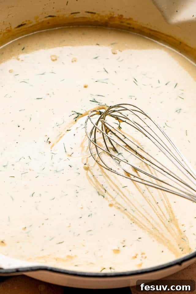 A hand whisking creamy dill sauce in a white pan, showing the rich, smooth texture of the simmering sauce.