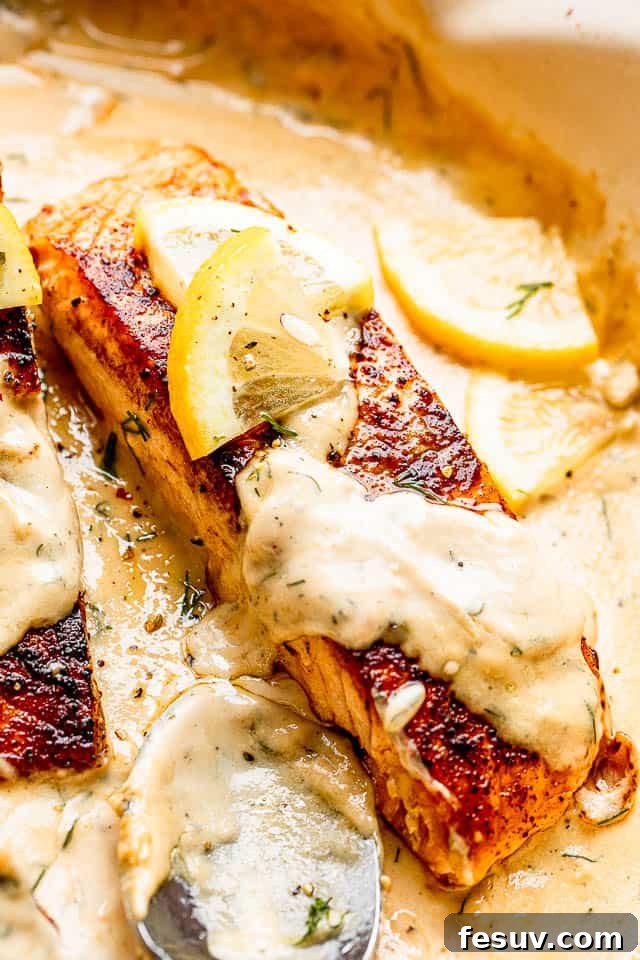 Close-up of a single cooked salmon fillet, flaking apart, topped with a generous dollop of creamy dill sauce and a fresh lemon slice.
